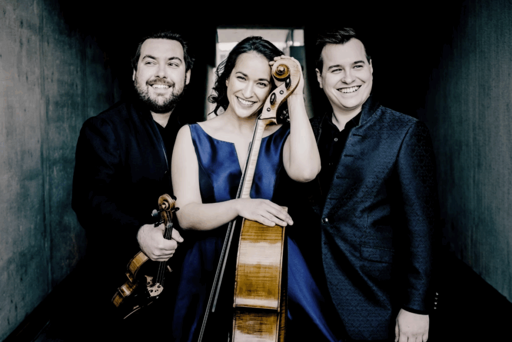 Drei Musiker:innen stehen nebeneinander in einem dunklen Durchgang. Die Frau in der Mitte trägt ein elegantes, dunkelblaues Kleid und hält ein Cello, auf dem sie sich anlehnt. Links von ihr steht ein Mann mit Bart, der eine Geige hält, und rechts ein weiterer Mann in einem dunkelblauen Anzug. Alle drei lächeln und blicken freundlich zur Kamera.
