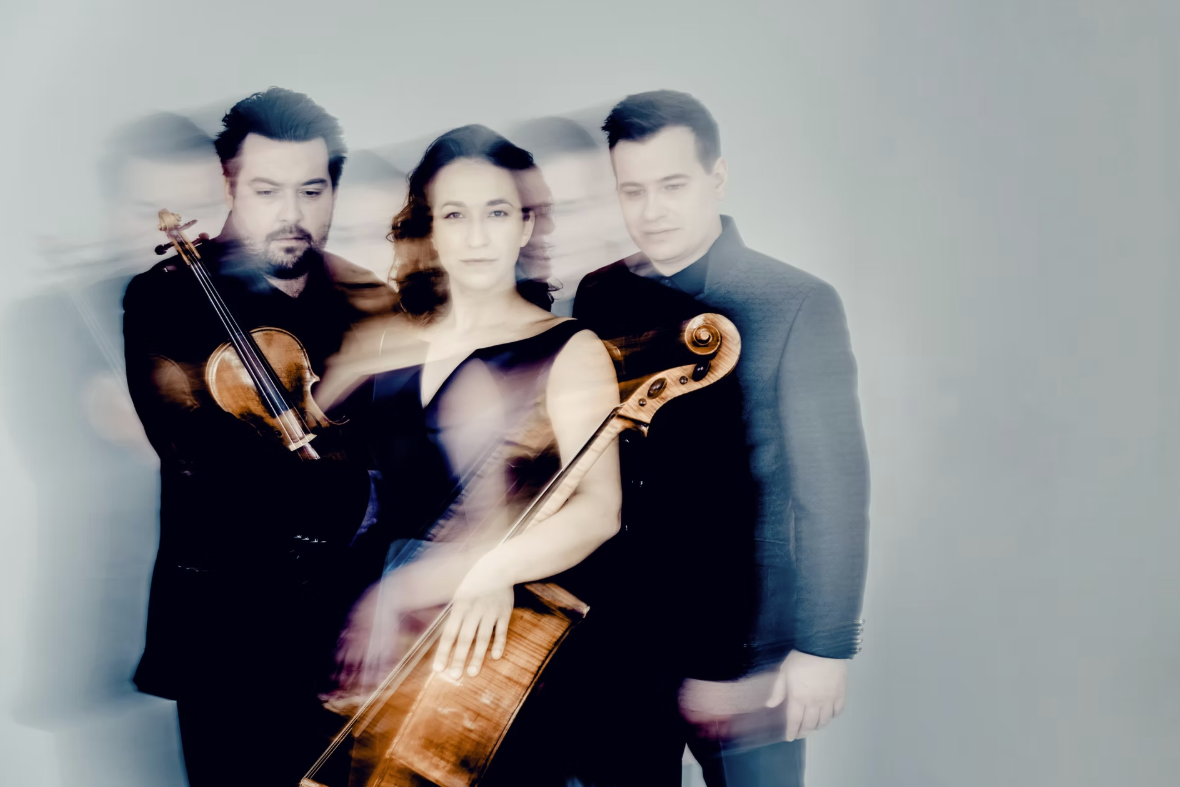 Drei Musiker:innen stehen vor einem hellen Hintergrund. Die Frau in der Mitte hält erneut ein Cello, während der Mann links eine Geige trägt. Das Bild hat einen künstlerischen Unschärfeeffekt, der eine Bewegungsdynamik erzeugt – insbesondere sind die Umrisse mehrfach zu sehen. Die Gesichter wirken ernst und konzentriert.